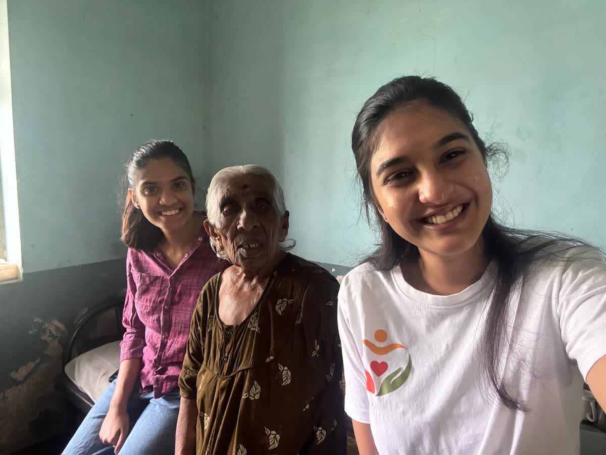 Two volunteers sitting on a cot beside an elderly resident, all three smiling at the camera