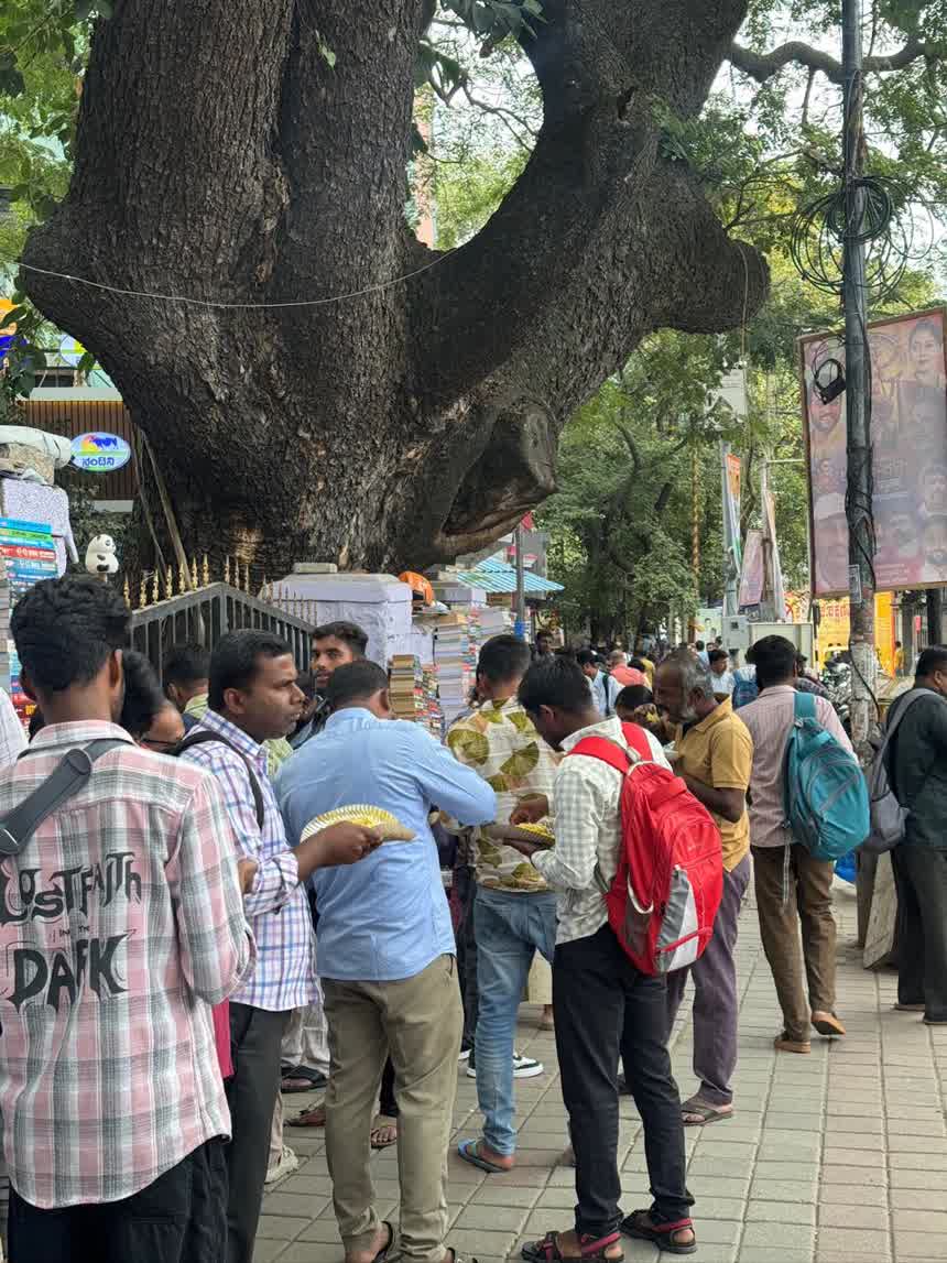 People standing on the footpath eating from paper plates under a large tree